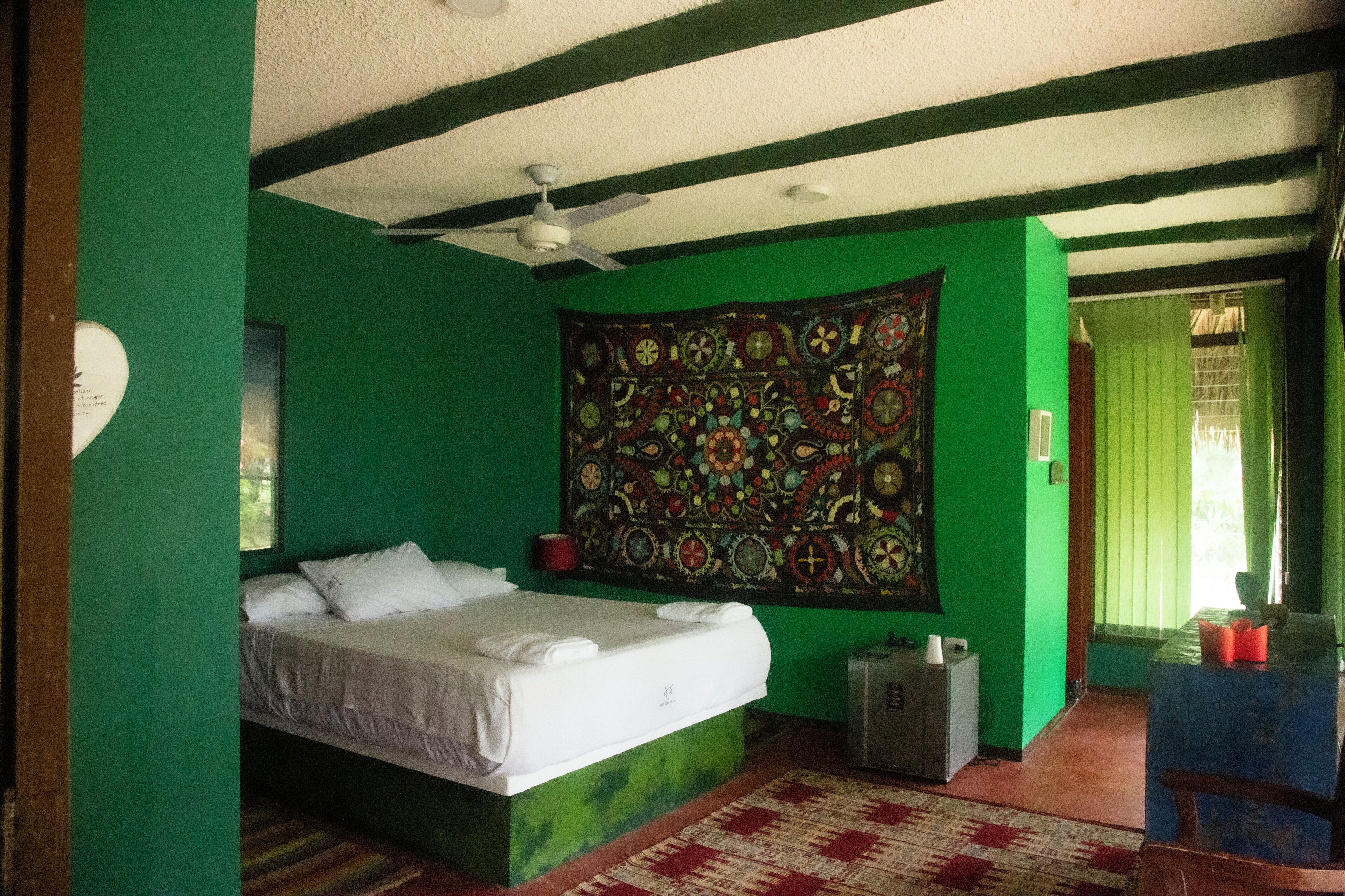 Una habitación con cama, un tapiz, ventilador de techo y una ventana con cortinas.