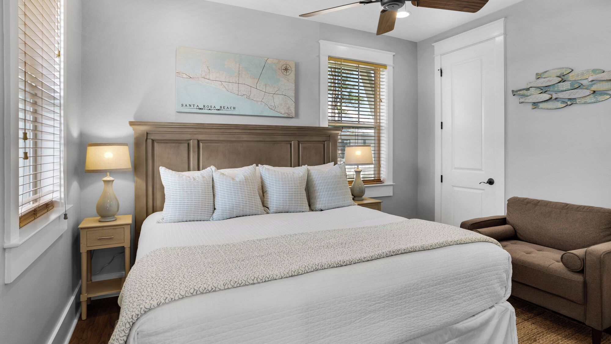 A bedroom with a large bed, two bedside tables with lamps, a ceiling fan, a map on the wall, and a door.
