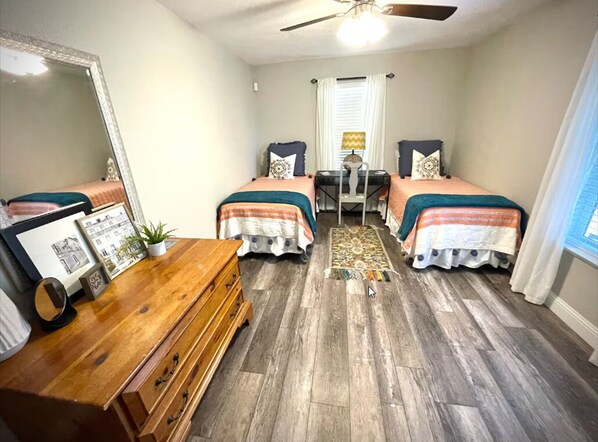 Une chambre avec deux lits, une commode en bois, un bureau, une chaise, un ventilateur de plafond et une fenêtre avec des rideaux.