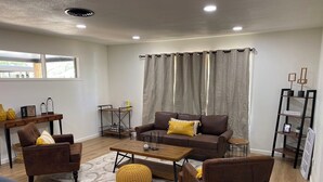 A modern living room with a sofa set, coffee table, and armchairs. There is a window with curtains, a small side table, and a bookshelf.