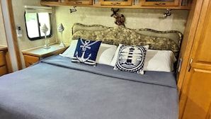 A bed with a grey textured bedspread, two decorative pillows with nautical designs, a nightstand with a lamp, and a window with a view of greenery. - Cozy 1-bedroom mobile home with AC, WiFi in enchanting Key Largo (Key Largo)