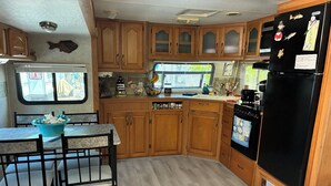 A compact RV kitchen with wooden cabinets, a black refrigerator, a stove, and a dining area with a table and chairs. - Cozy 1-bedroom mobile home with AC, WiFi in enchanting Key Largo (Key Largo)