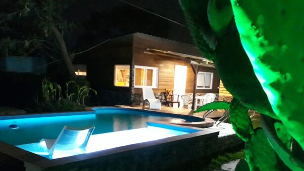A nighttime view of a house with a swimming pool, a patio with chairs, and a well-lit interior. - Vivisierras Espectacular Cabaña Villa Carlos Paz pileta privada barrio exquisito (Villa Carlos Paz)