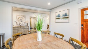 Un comedor con una mesa redonda de madera, seis sillas, un jarrón blanco con plantas verdes, un cuadro en la pared y una puerta de madera.