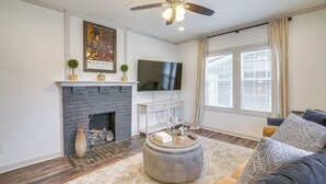 Una sala de estar con chimenea, un televisor montado encima, un ventilador de techo y un ventanal con cortinas.