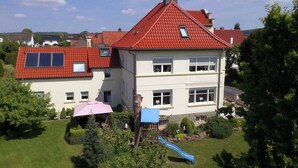 Een huis met twee verdiepingen, een rode pannendak, een blauwe glijbaan en een roze parasol in de tuin.