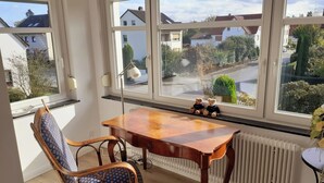 Een kamer met een houten tafel, twee stoelen en een raam met uitzicht op huizen en groen.