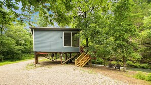 A small cabin with a wooden staircase and a bench near a river, surrounded by trees. - Pam's Treehouse Cabin #2-Your wonderful treehouse getaway...right on a creek! (Hot Springs)