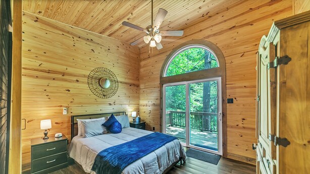 A wooden-paneled bedroom with a bed, nightstands, a ceiling fan, and a large arched window. - Pam's Treehouse Cabin #2-Your wonderful treehouse getaway...right on a creek! (Hot Springs)