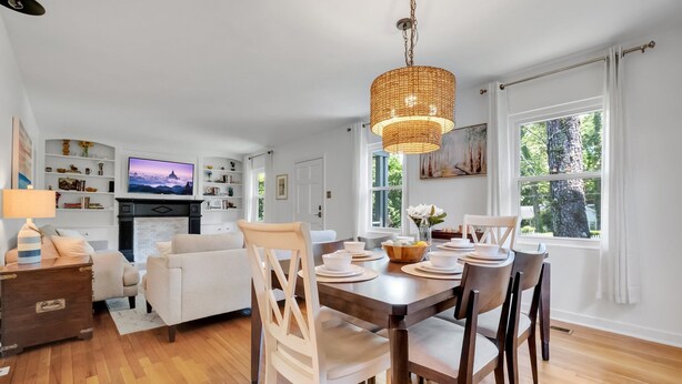 A modern living room with a dining area, wooden flooring, white furniture, a television, and a large window with a view of trees. - Charming 3/2 Midtown Home. Minutes from FSU. Fenced Backyard. Pet-Friendly. (Tallahassee)