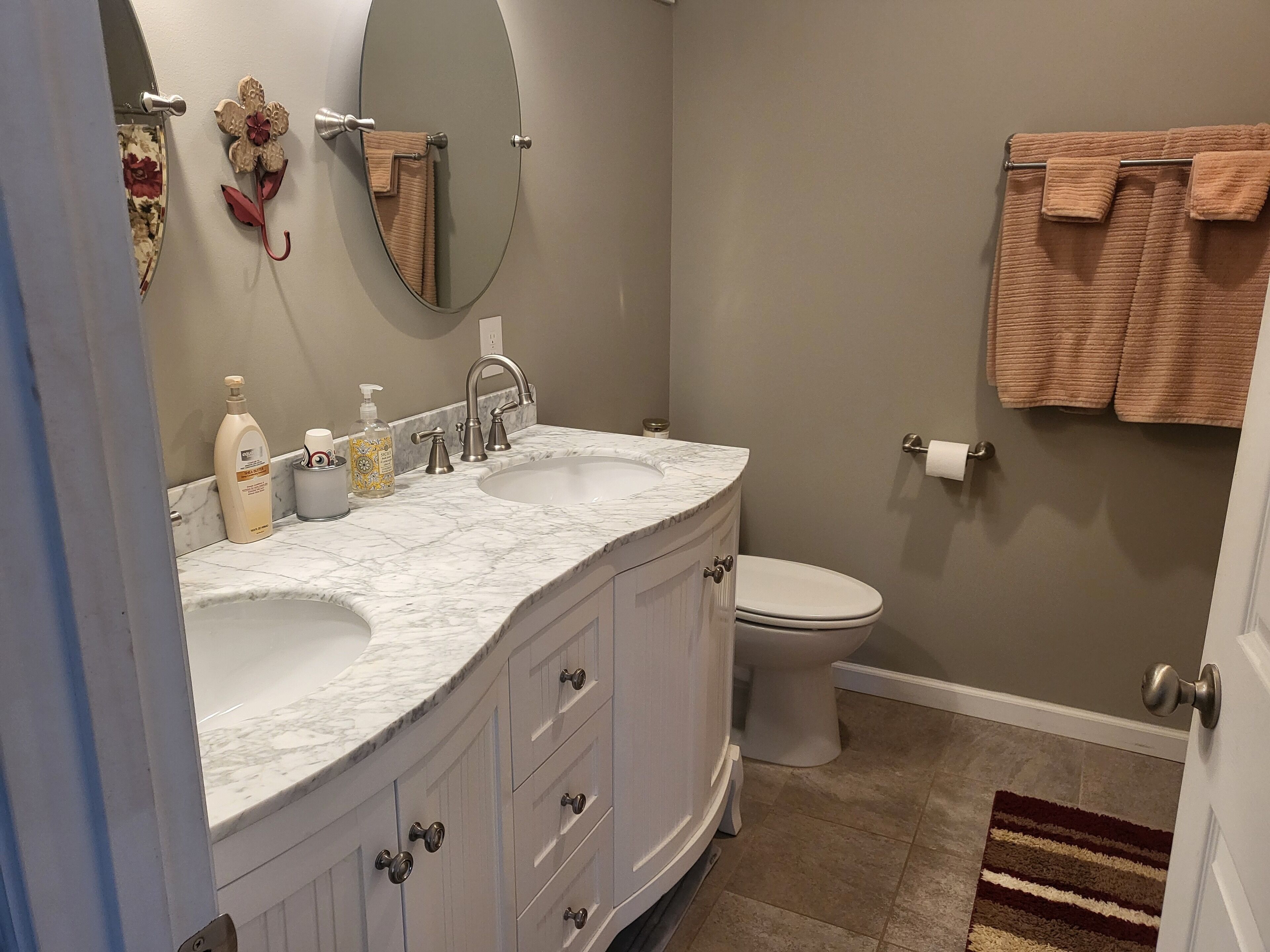 A bathroom with a double sink vanity, a toilet, a towel rack, and a decorative flower on the wall.