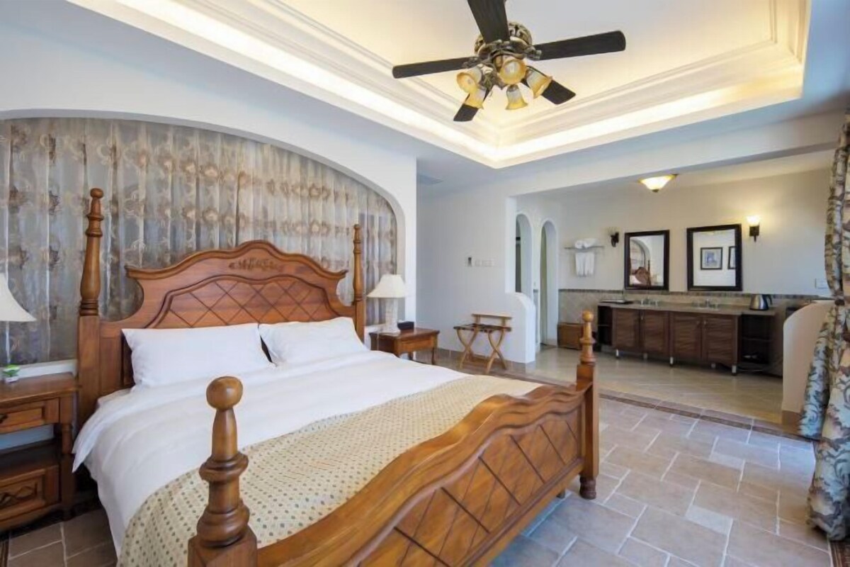 Une chambre spacieuse avec un grand lit, un ventilateur de plafond et une vue sur la salle de bain et la cuisine.