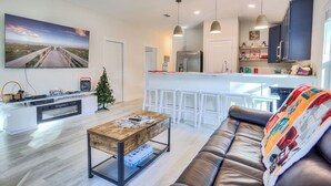 A modern living room with a leather sofa, a coffee table, a TV, and a kitchen area with white bar stools.