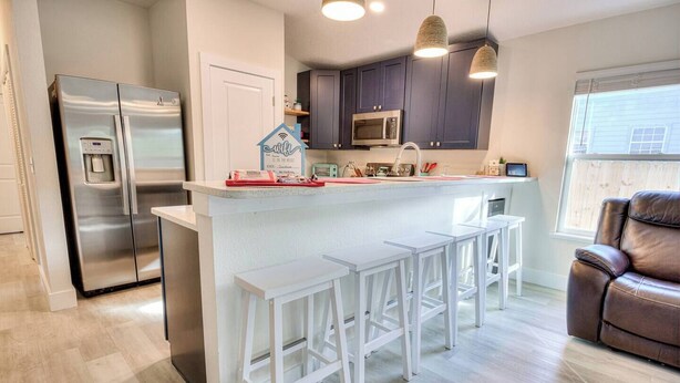 A modern kitchen with stainless steel appliances, a white island with bar stools, dark cabinets, and a large window.