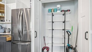 A compact kitchen with a stainless steel refrigerator, a built-in shelf with cleaning supplies, and a broom leaning against the wall.