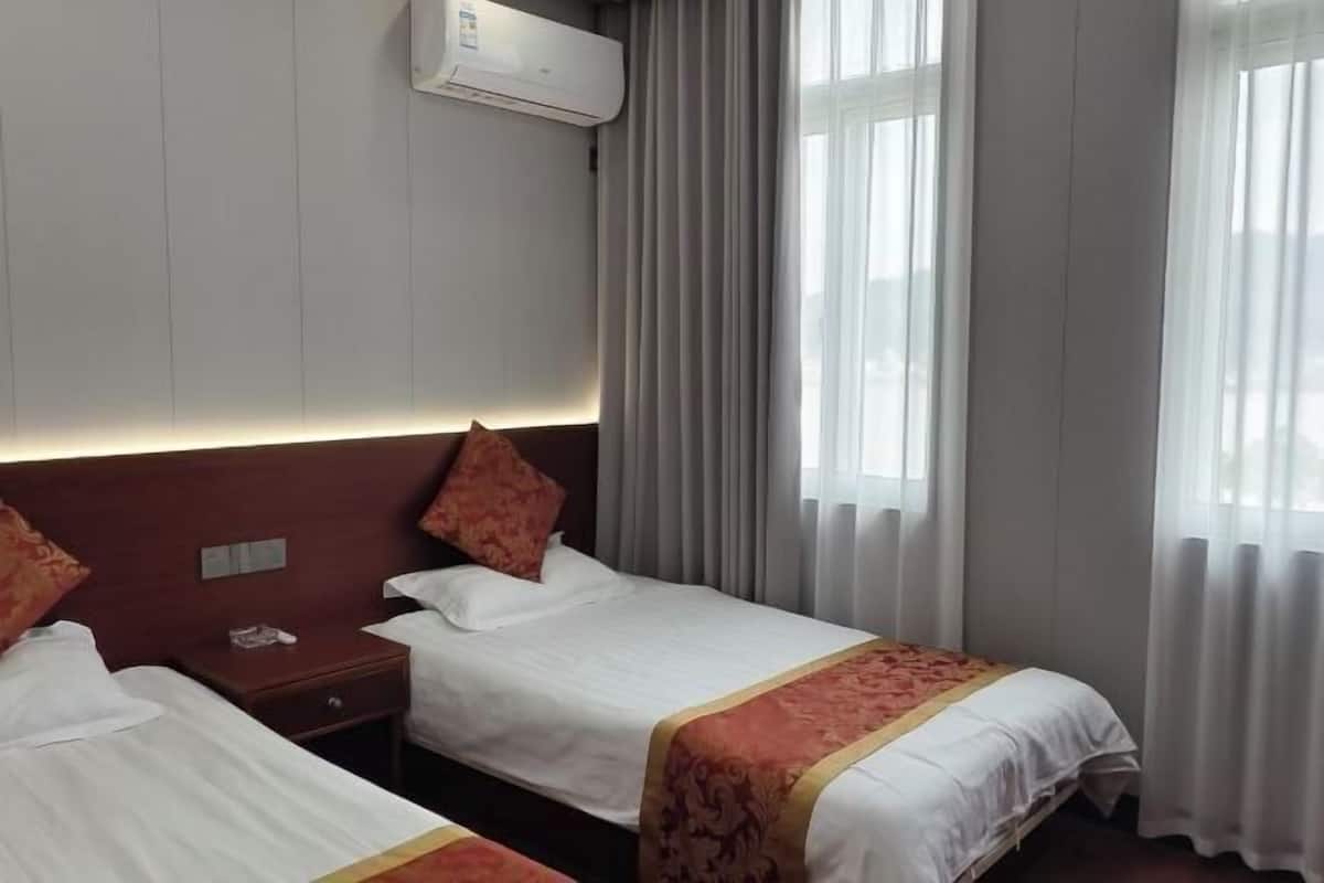 A hotel room with two beds, a nightstand, an air conditioning unit, and a window with curtains.