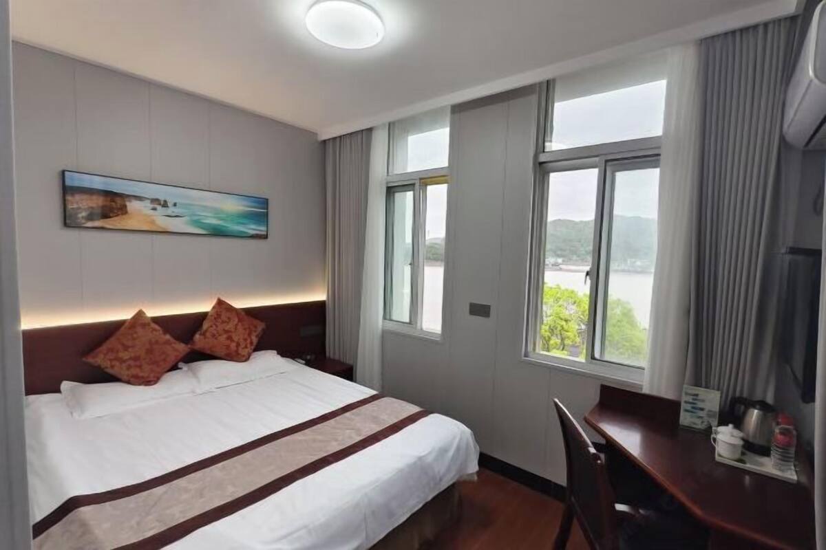 A hotel room with a bed, a desk, a painting, and a window with a view.