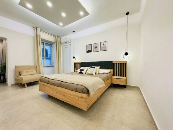 La stanza è dominata da un elegante letto di design con struttura in legno e rivestimento neutro. Completo di lenzuola bianche e coperte morbide, è affiancato da lampade a sospensione moderne e un comodino minimalista.