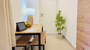 Une petite pièce, bien rangée, avec une table en bois, un ordinateur portable, un panier de fruits, une lampe et une plante en pot.