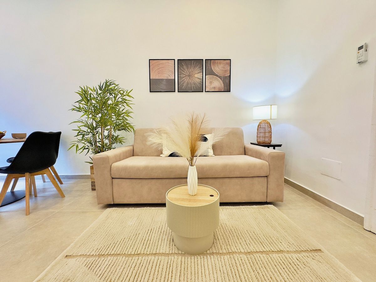 A living room with a beige sofa, a round coffee table with a vase, a potted plant, and a wall-mounted lamp.