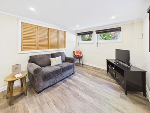 A living room with a grey sofa, a small wooden table, a TV stand, and a window with blinds.
