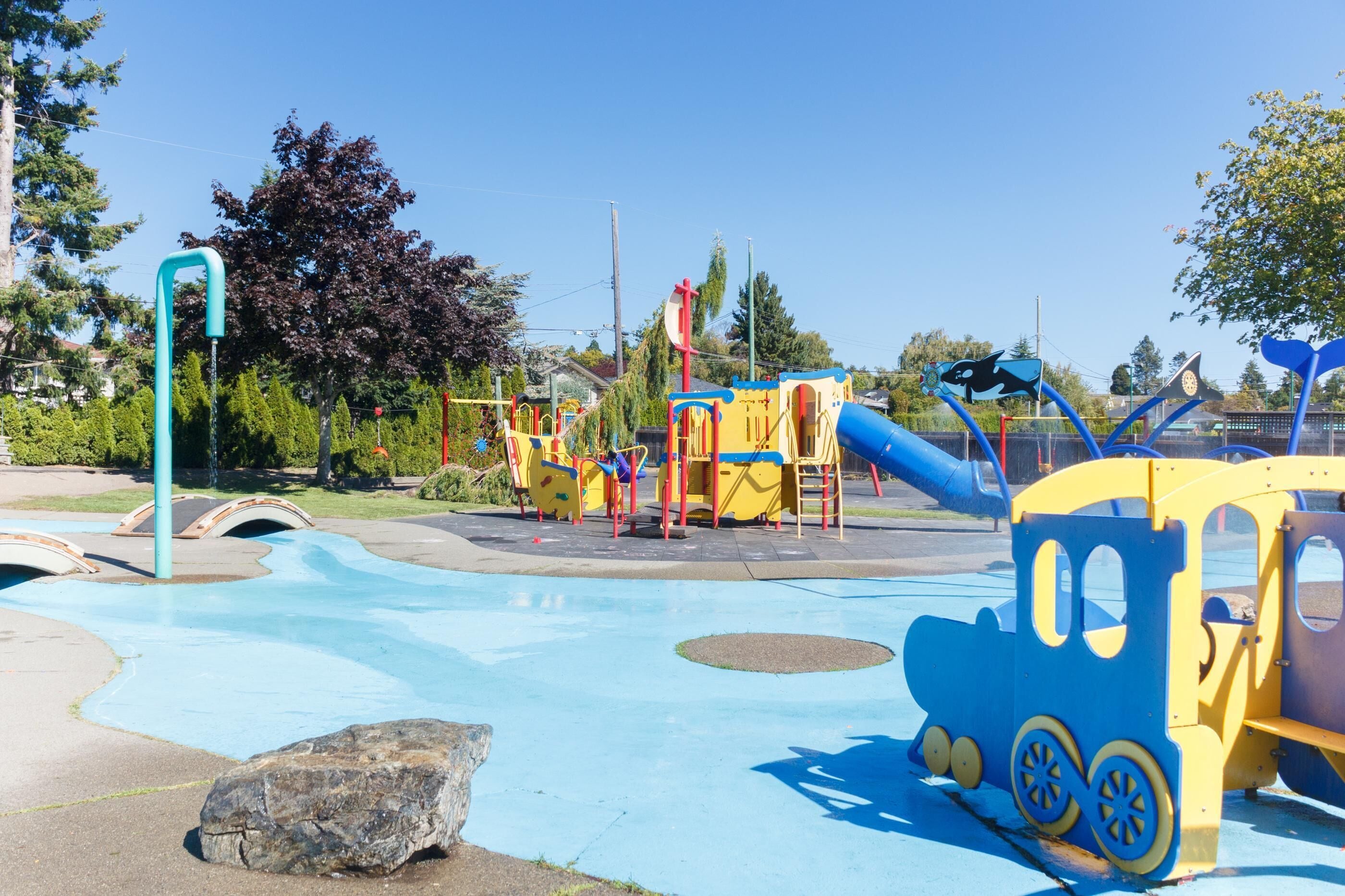 A playground with a blue and yellow train-themed play structure, a water play area, and a large rock in the foreground.