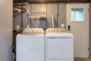 Two white front-loading washers in a laundry room with visible plumbing and a small window. - The Forrester Grove (Oak Bay North)