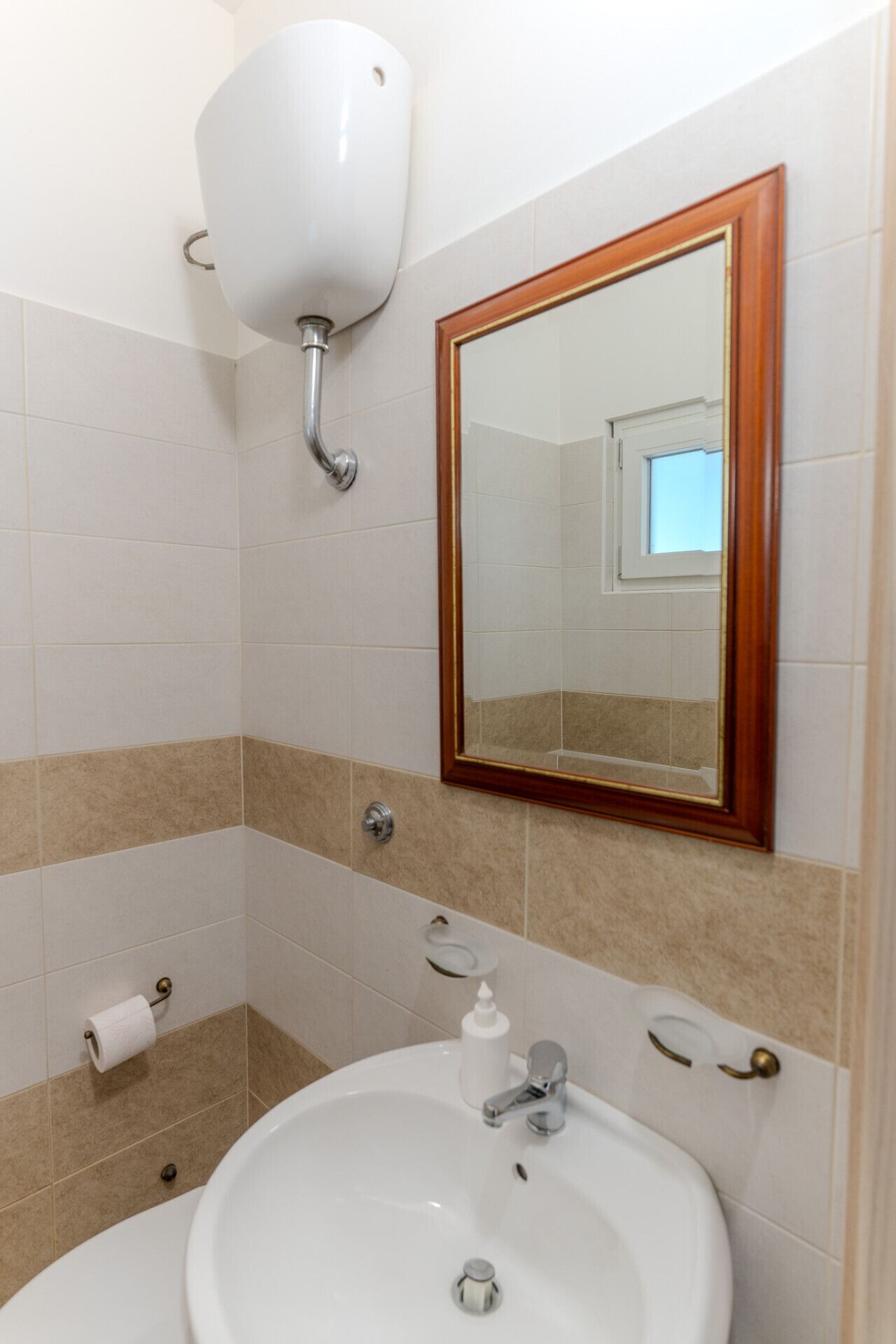 Une salle de bain avec un lavabo blanc, un miroir avec un cadre en bois, un porte-rouleau de papier toilette et un distributeur de savon.