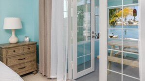 Une chambre avec une table de chevet en bois, une lampe blanche et une horloge. Une porte donne sur un balcon d’où l’on peut admirer la mer et les bateaux.