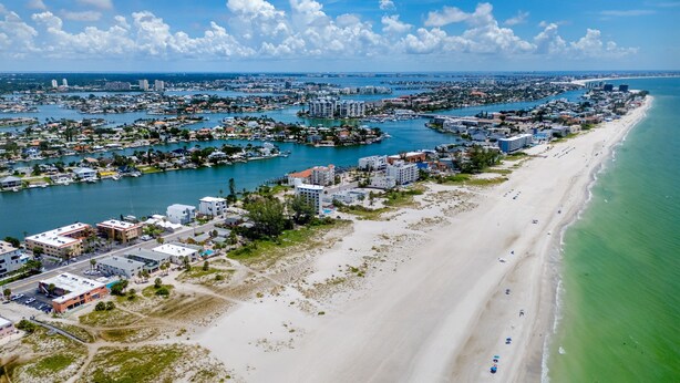 Vue aérienne d’une ville côtière avec une longue plage de sable, un port de plaisance et une zone urbaine dense, avec des bâtiments et des bateaux.