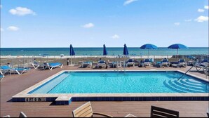 A pool area with a view of the ocean, blue umbrellas, and lounge chairs.