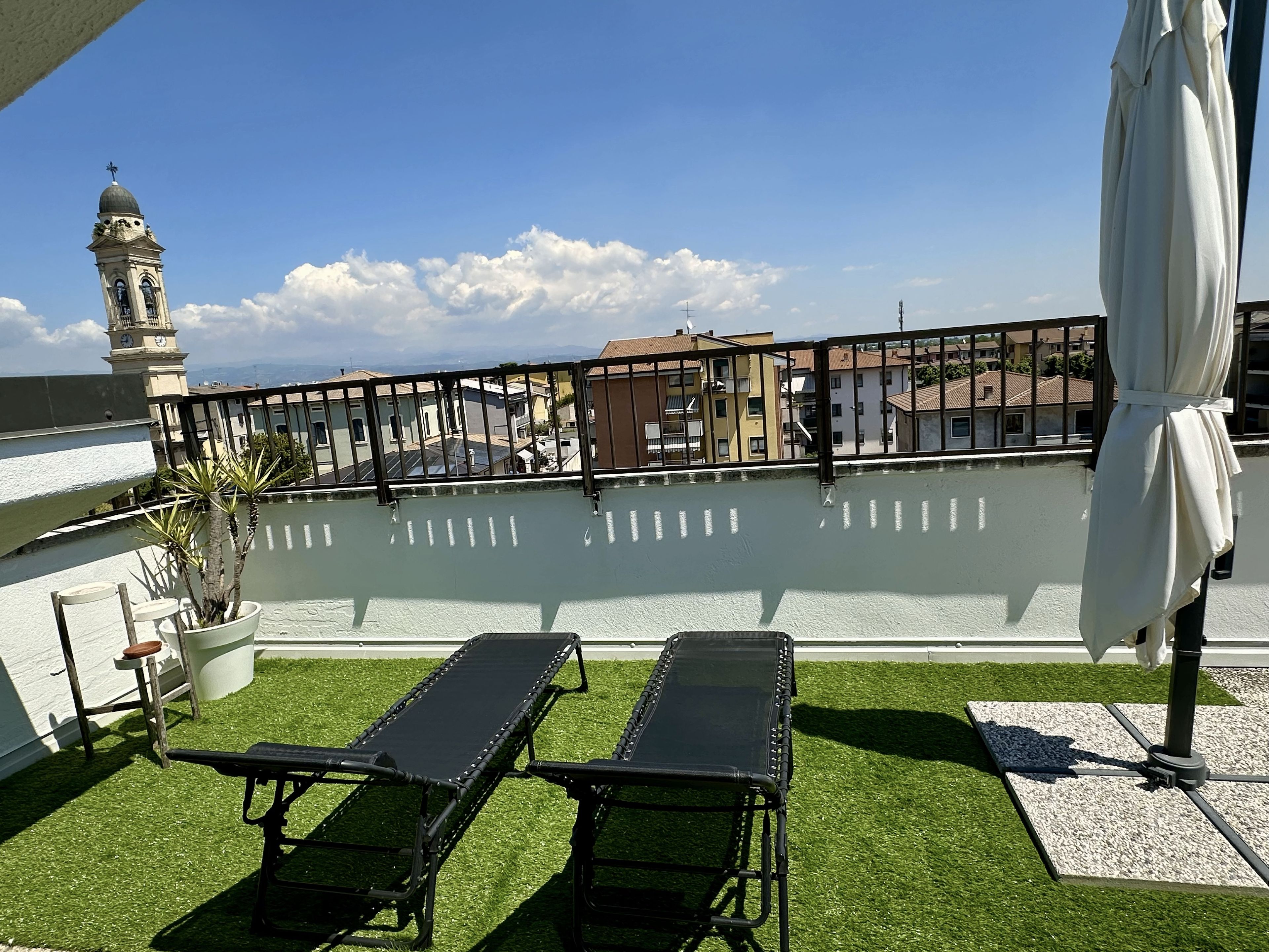 A rooftop terrace with two lounge chairs, a potted plant, and a view of buildings and a clock tower.