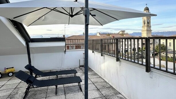 A balcony with two lounge chairs and a large umbrella, overlooking a cityscape with a prominent tower. - Catullo Loft (Dossobuono)
