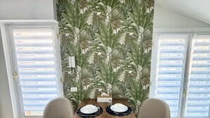 A dining area with a wooden table set for two, flanked by two chairs. The room features a large window with blinds, a tropical leaf-patterned wall, and a woven rug on the floor. - Catullo Loft (Dossobuono)