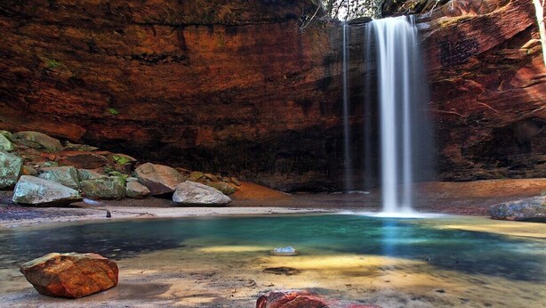 Une piscine naturelle formée dans le roc, avec une cascade, entourée de gros rochers et d’une formation rocheuse ressemblant à une grotte.