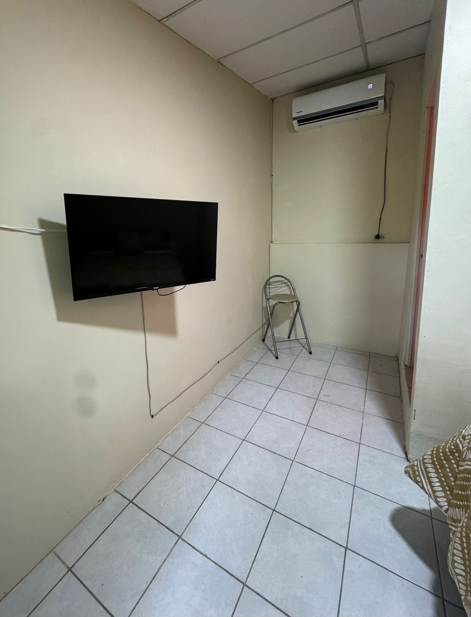 Una habitación con televisión de pantalla plana montada en la pared, una pequeña silla plegable y un equipo de aire acondicionado en el techo.