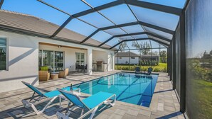 A covered outdoor swimming pool with lounge chairs, a patio area with outdoor furniture, and a view of a neighboring house.