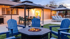 Eine Terrasse im Freien mit Feuerstelle, blauen Adirondack-Stühlen, einem Esstisch und einem überdachten Sitzbereich mit Lichterketten.
