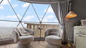 Ein Zimmer mit großem Fenster, zwei Sesseln, einem kleinen Tisch, einer Lampe und einem altmodischen Schrank.