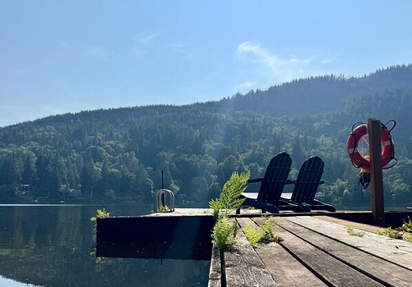 Un muelle de madera con dos sillas Adirondack, un flotador salvavidas y una pequeña estructura sobre un lago, con colinas boscosas al fondo.