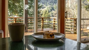 A dining table with a plate of food and a cup, overlooking a wooden deck and forested hills.