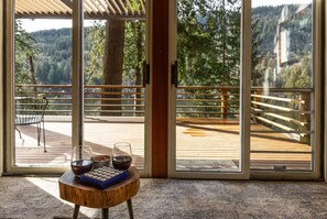 A room with a view of a wooden deck and forest. There is a table with a board game and glasses. - Lakeside Cabin w/ Dock, Boats & Stunning Views (Sedro-Woolley)