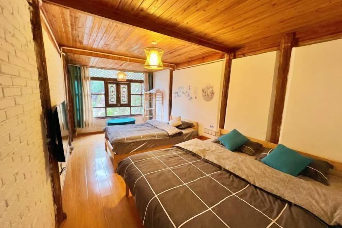 A room with wooden ceiling beams, two beds with grey and white bedding, a window with curtains, and a hanging light fixture.