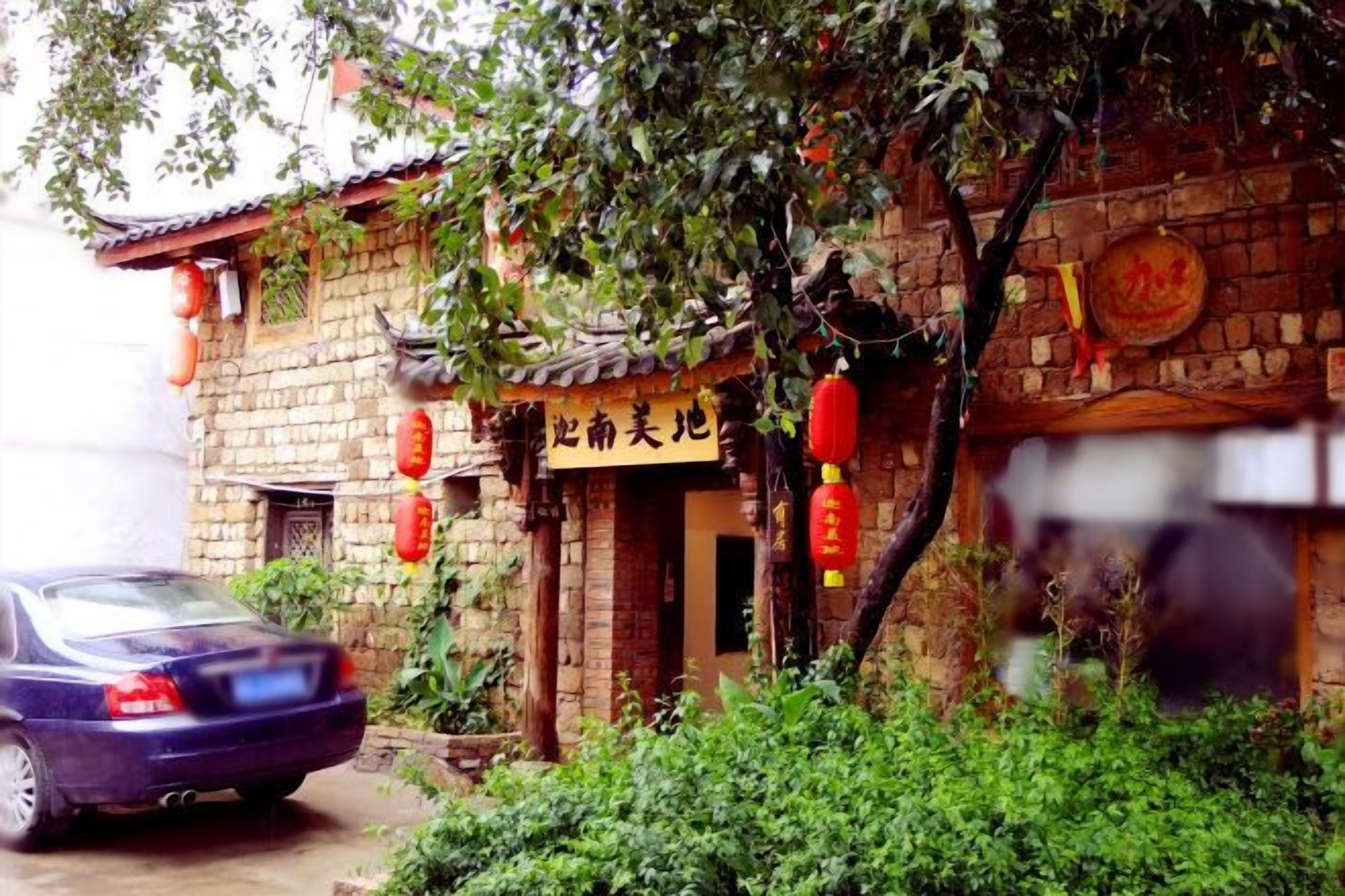 A traditional brick building with a signboard, a parked car, and a tree in the foreground.