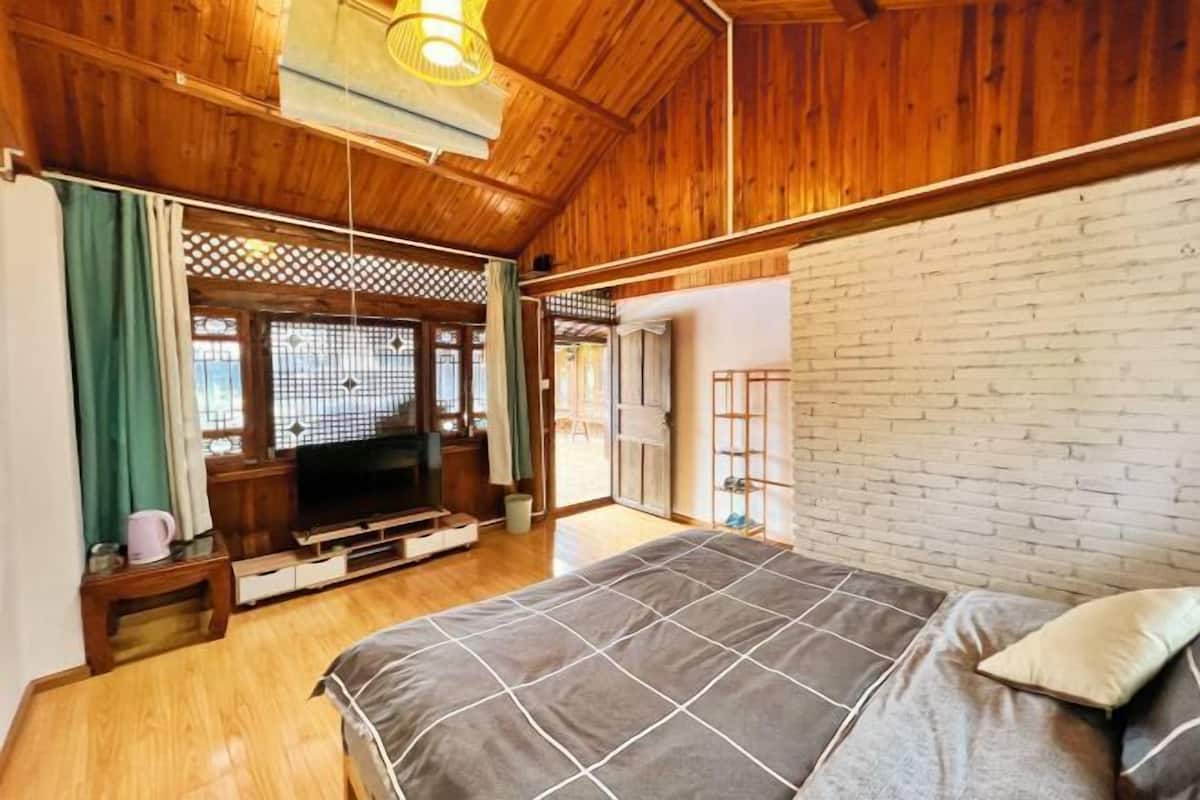 A bedroom with a wooden ceiling, a brick wall, a bed with a grey quilt, a television on a wooden stand, and a sliding door leading to another room.