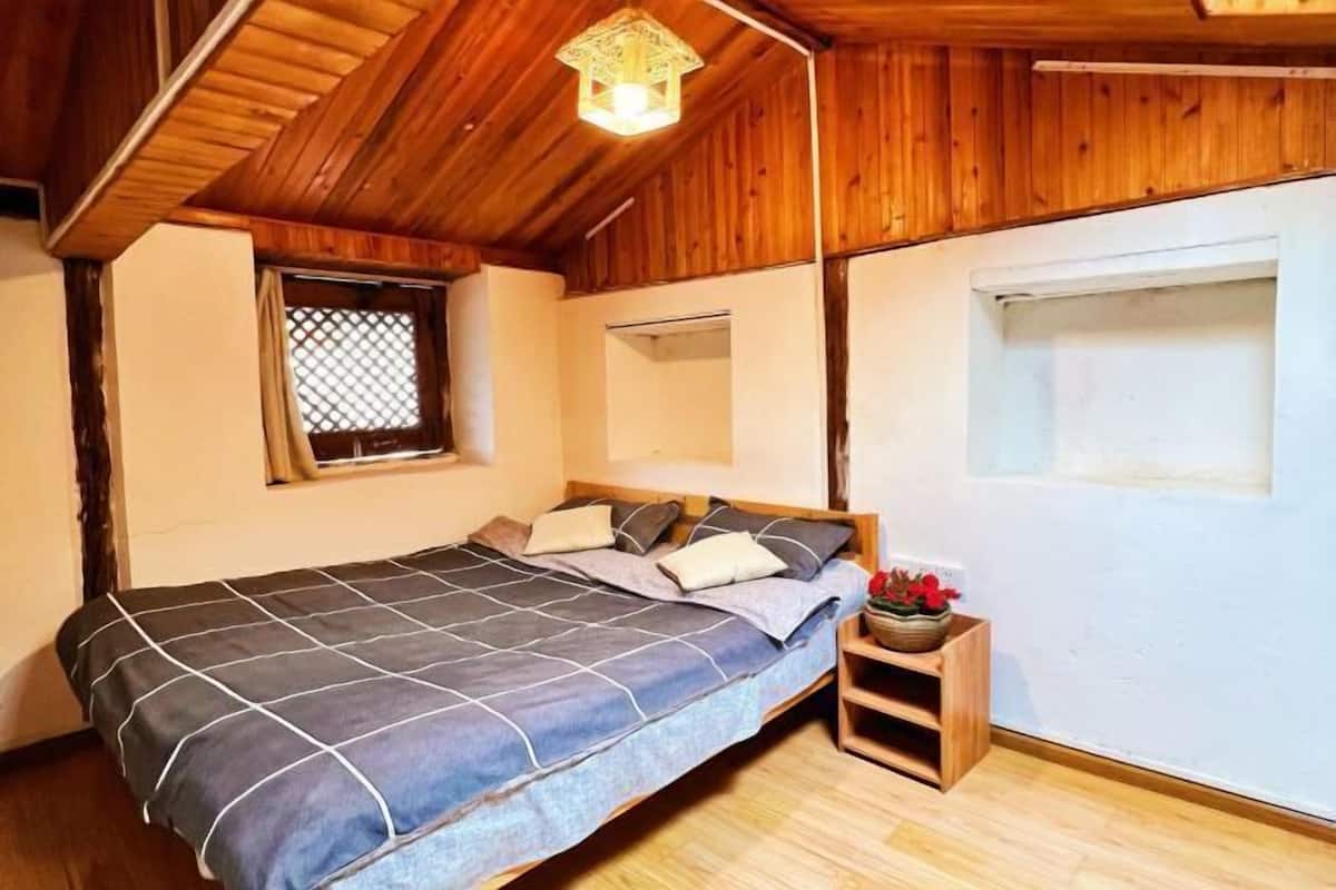 A bedroom with a wooden ceiling, a bed with a blue and white bedspread, a small wooden nightstand with a flower vase, and a window with a lattice design.