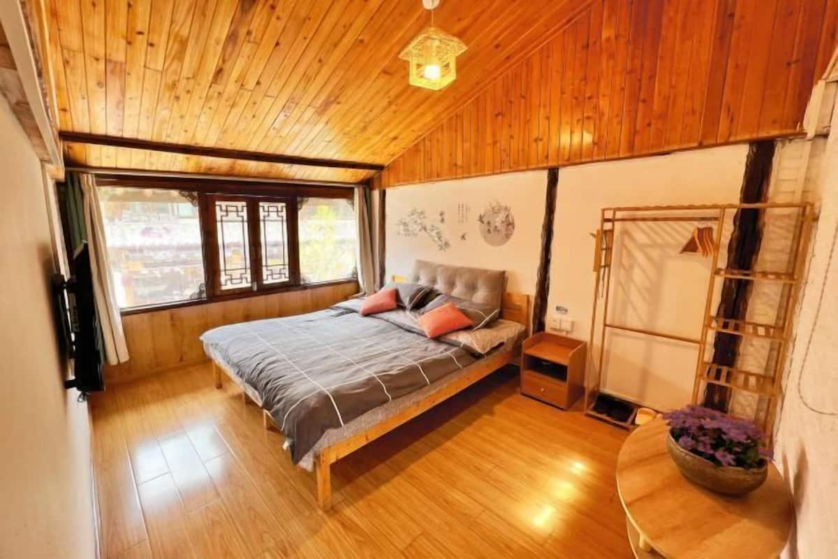 A wooden-paneled room with a bed, a window, a wall-mounted shelf, and a small table with a flower arrangement.