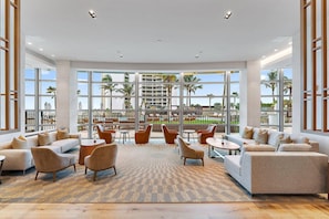 A spacious lounge area with multiple seating options, large windows offering a view of a pool and palm trees, and a modern ceiling design.
