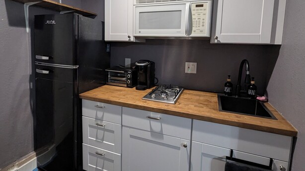 Cocina compacta con gabinetes blancos, refrigerador negro, microondas, cafetera y lavamanos con dispensadores de jabón.
