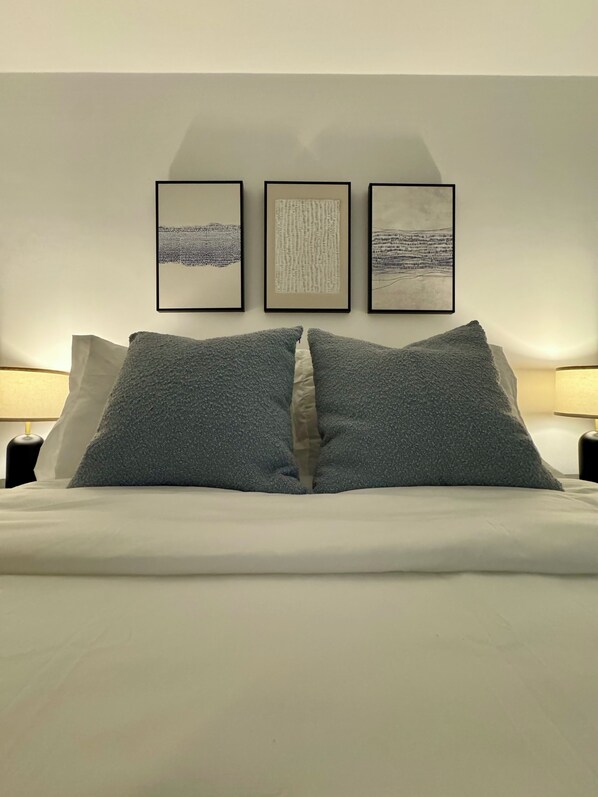 Un lit bien fait, avec deux oreillers gris et une couette blanche, flanqué de deux tables de chevet sur lesquelles reposent des lampes. Trois œuvres d’art encadrées sont accrochées au mur au-dessus du lit.